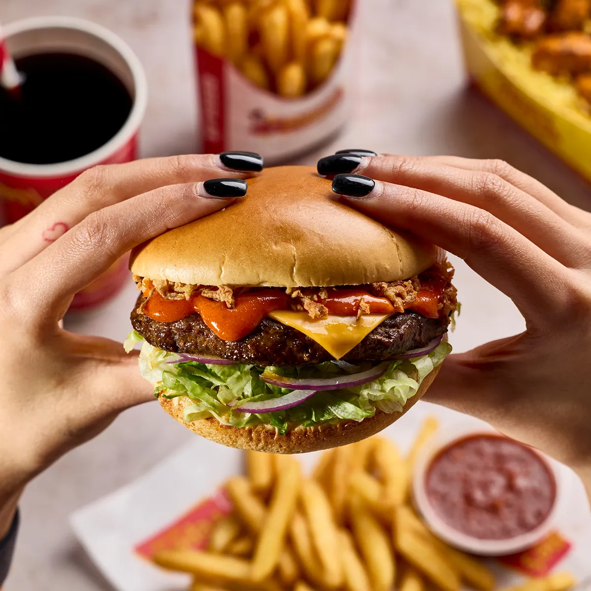 A delicious burger being held in 2 hands with some chips and a soft drink in the background.
