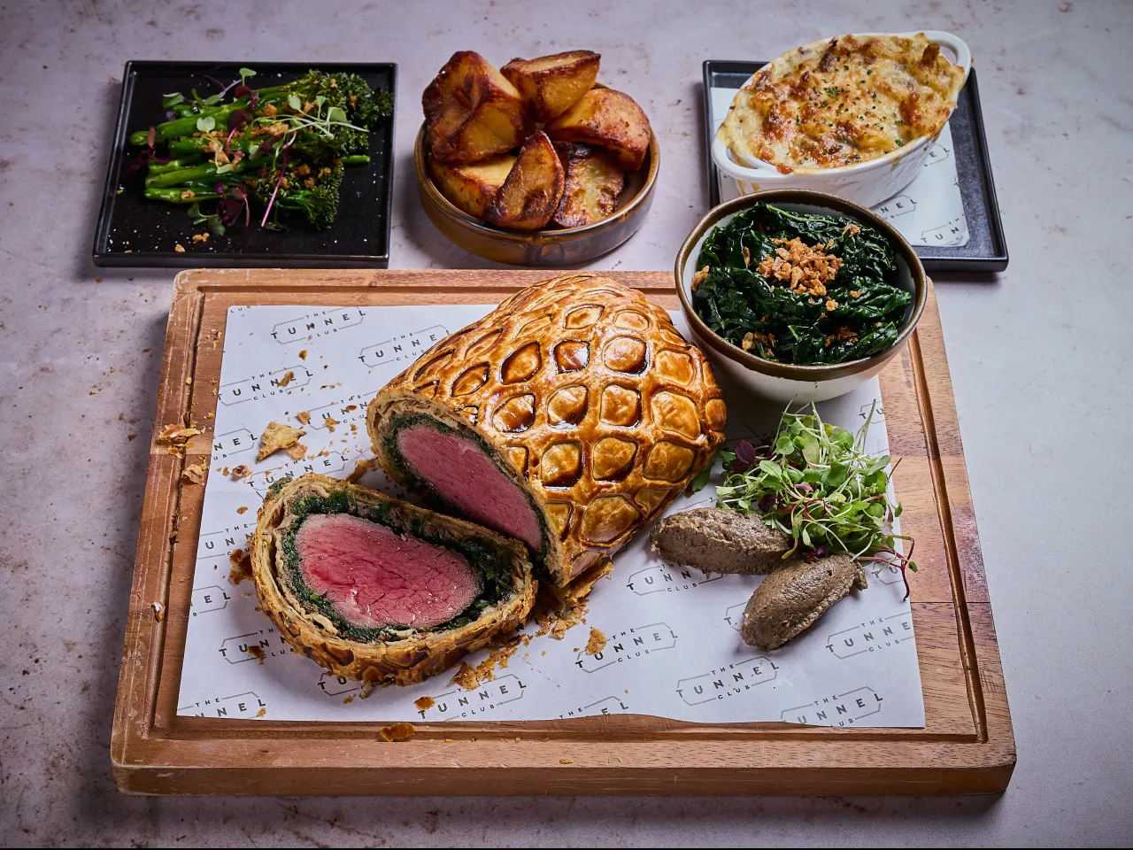 A beef wellington sliced with side dishes on a wooden chopping board.