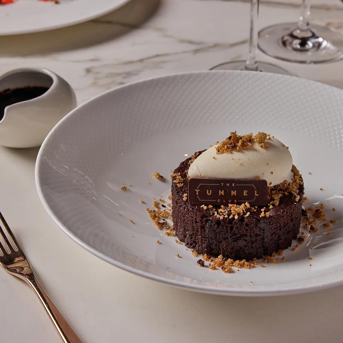 A dessert in a bowl with cutlery, sauce and glasswear on a table.