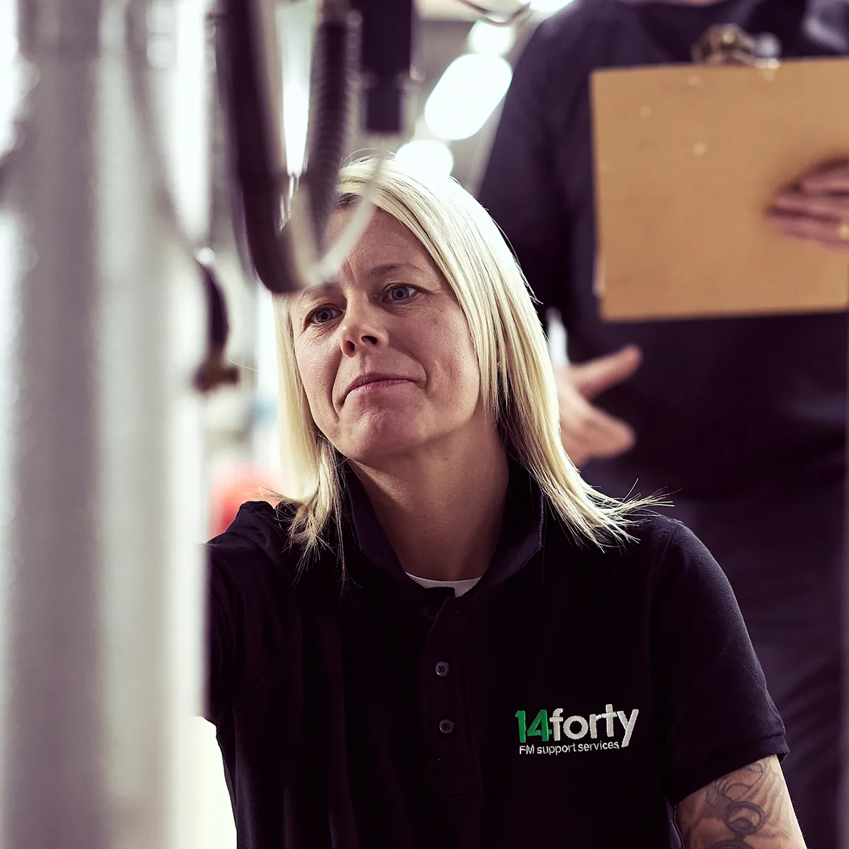 A female looking into the distance within a work environment with someone with a clipboard behind her.
