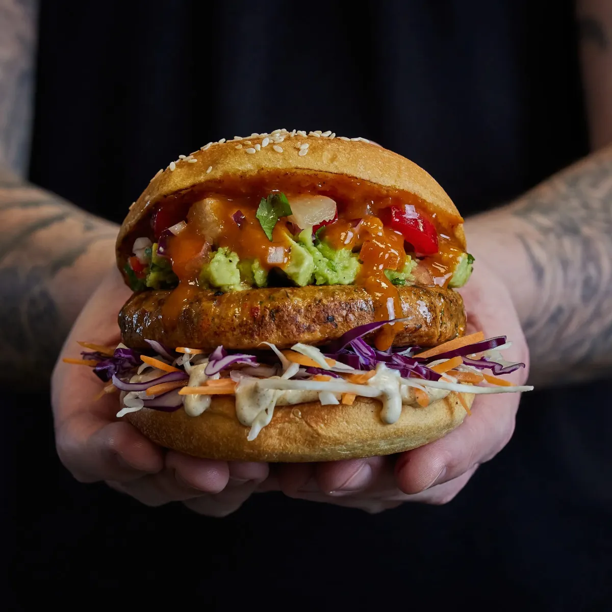 A chef holding a delicious veggie burger with coleslaw, lettuce and sauce in a toasted sesame seed bun.