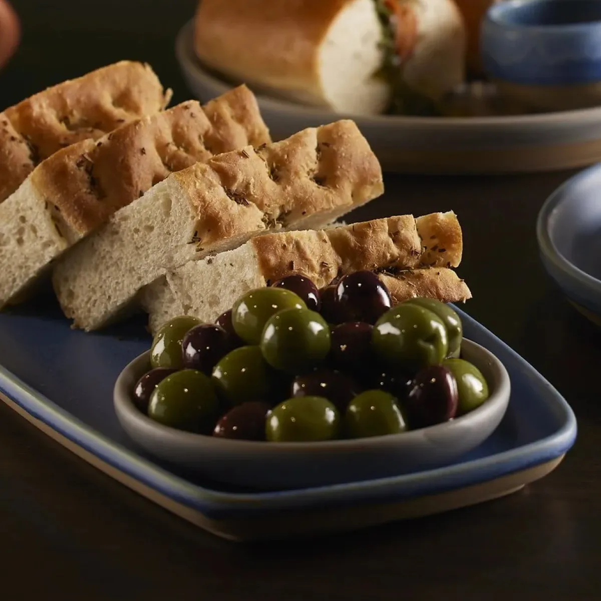 A bowl of olives with 2 different types of bread in the background.
