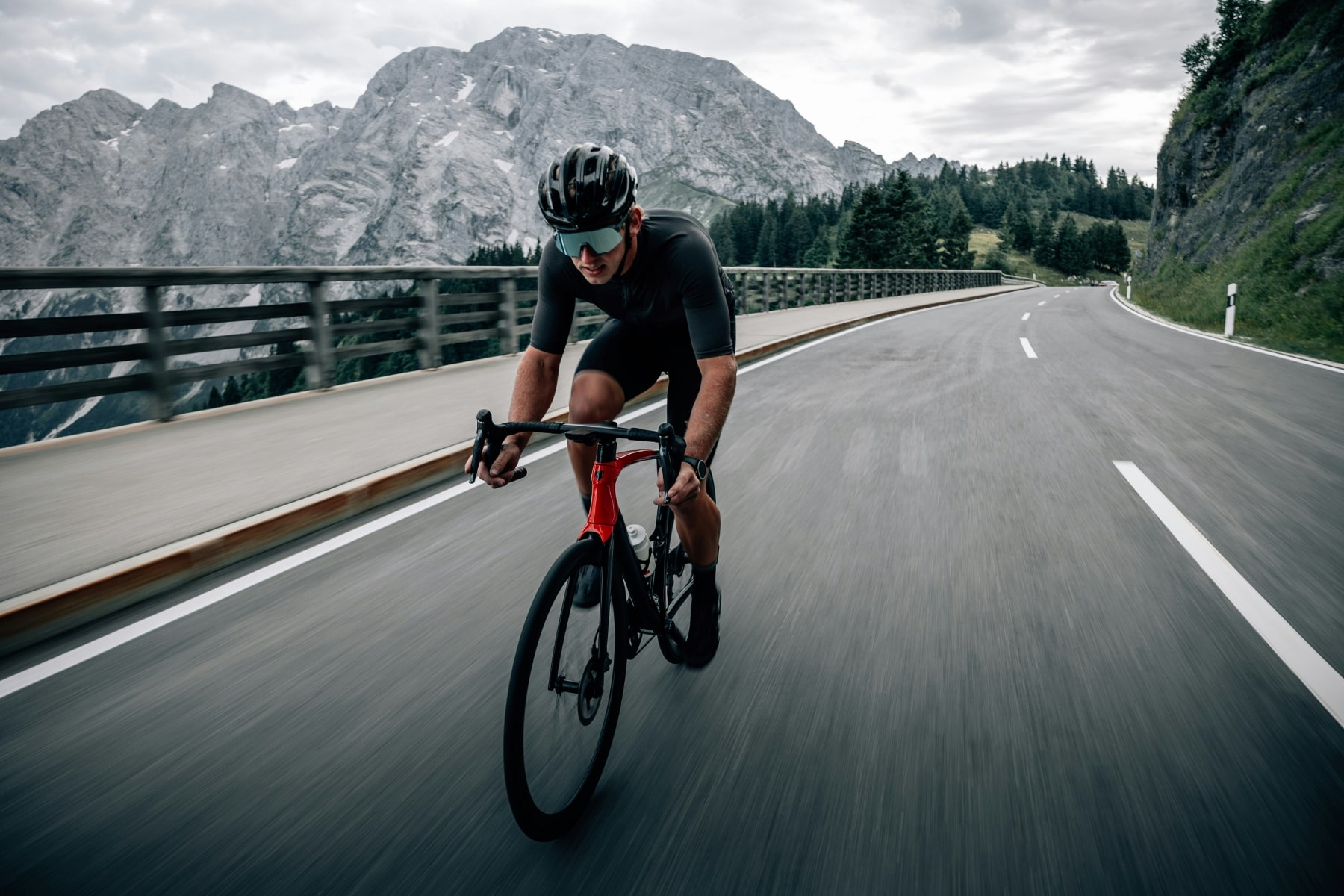 A cyclist riding fast on a winding mountain road with rocky peaks in the background.