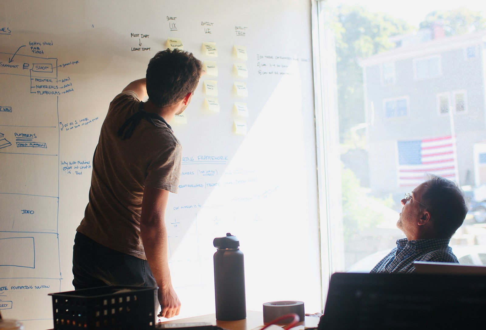 Two people standing at a whiteboard covered in notes while another person sits nearby in a sunlit office.