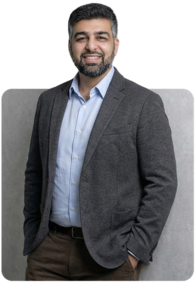 Smiling man with dark hair and beard wearing a grey blazer over a light blue shirt, standing with hands in pockets.