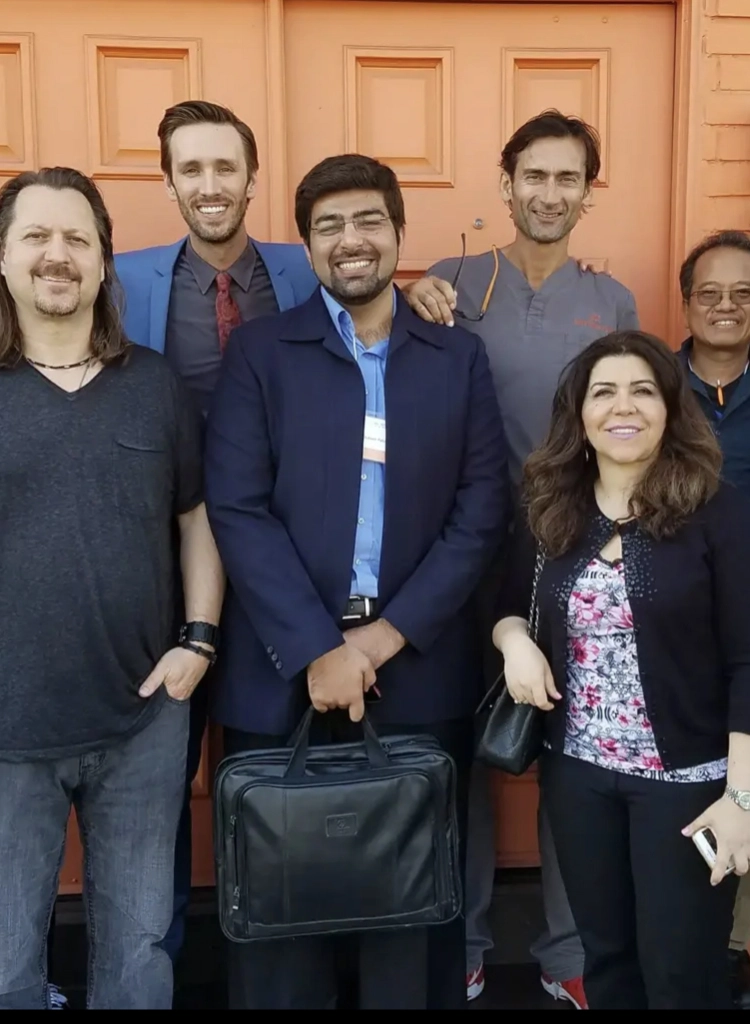 Group of six diverse adults smiling and posing in front of an orange door, one man holding a black briefcase.