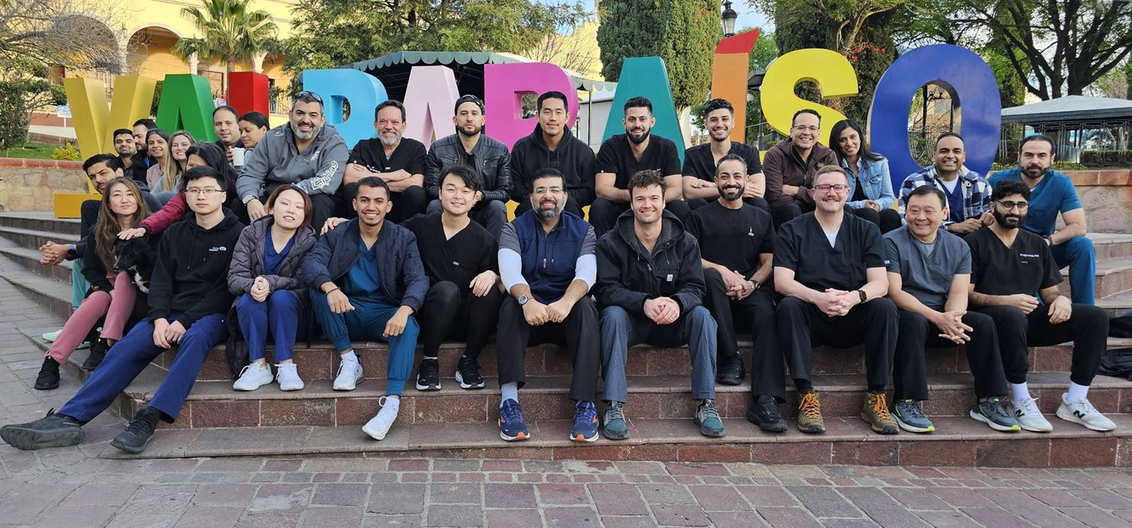 Large diverse group of people sitting on steps in front of colorful 'Valparaíso' sign outdoors.