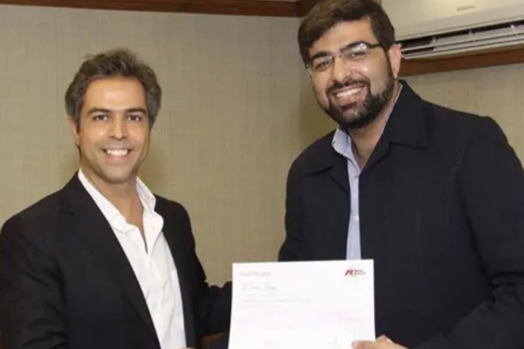 Two men smiling, one handing a certificate to the other indoors.