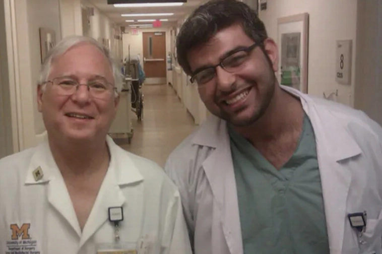Two smiling male medical professionals in a hospital corridor, wearing white coats and scrubs.
