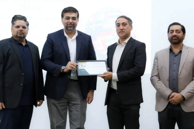 Four men in business attire standing against a white background, two men in the center holding a certificate together.