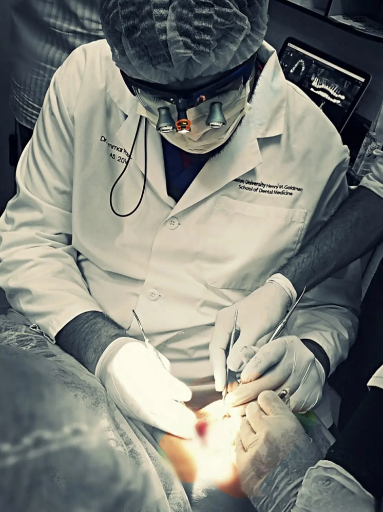 Dentist in protective gear performing a dental procedure on a patient with dental tools and a bright light.