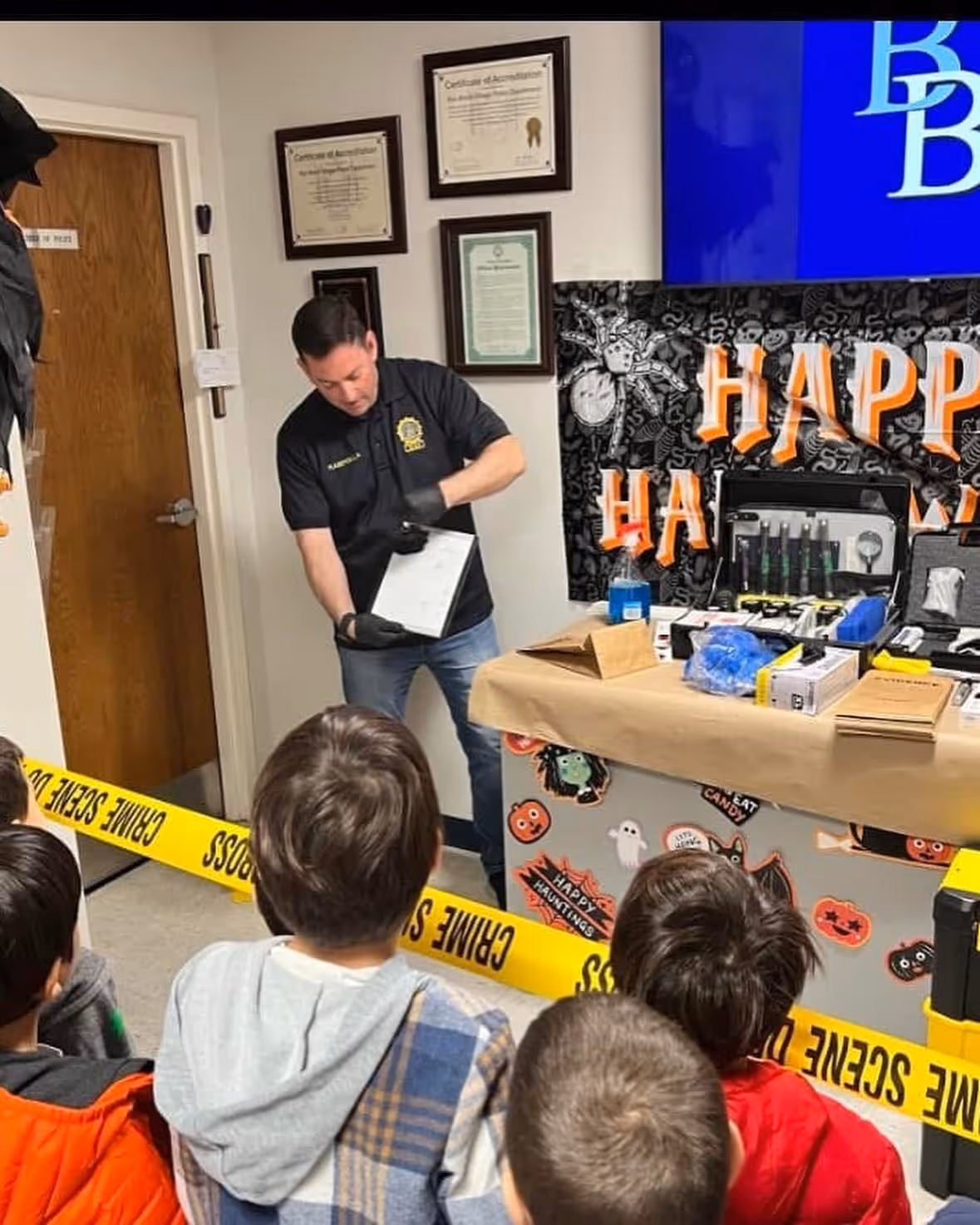 A man standing in a room with a group of kids, wearing jeans and casual clothing. The walls have text related to a "crime scene." The setting appears to be an indoor space resembling a shop or play area.
