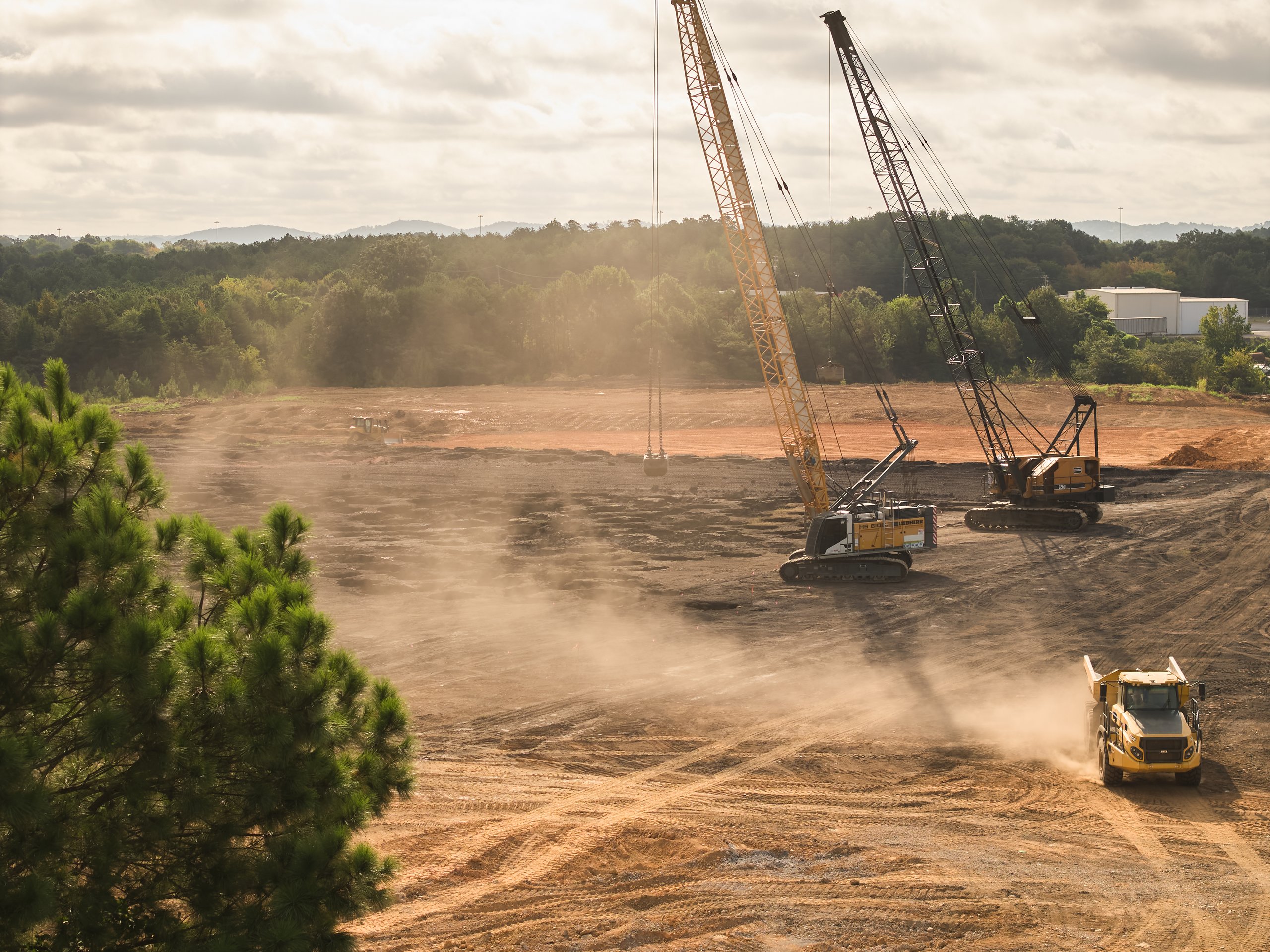 Dust rising behind a haul truck as two cranes compact the Coalburg site, seen past a pine, photographed by Omar Mohammad Studio