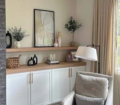 Cozy living room corner with a beige armchair, floor lamp, white cabinets, wooden shelves, decorative vases, framed photos, and a window with beige curtains.