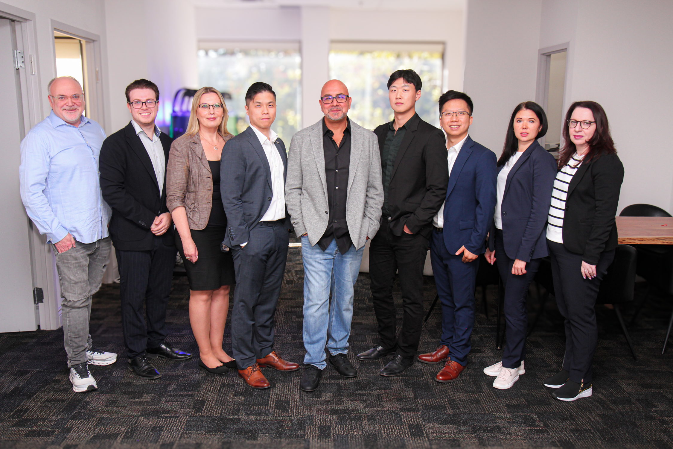 A group of nine team members posing in a pyramid formation. A man stands at the center while the others, six men and three women in total, stand and sit around him, looking toward the camera with smiles.