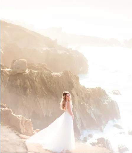 A woman in a wedding dress is standing in front of the sea on the rocks.