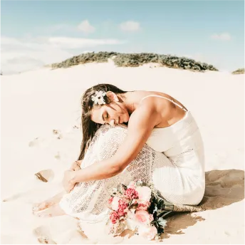 A woman in her wedding dress is sitting on the beach with flowers.