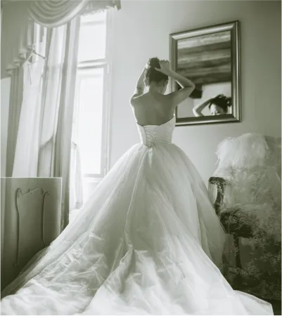 Black and white photo of a woman in a wedding dress in a room in front of a mirror from the back.
