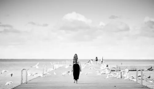 A woman stands in the middle of the pier, birds around her, she goes to the sea