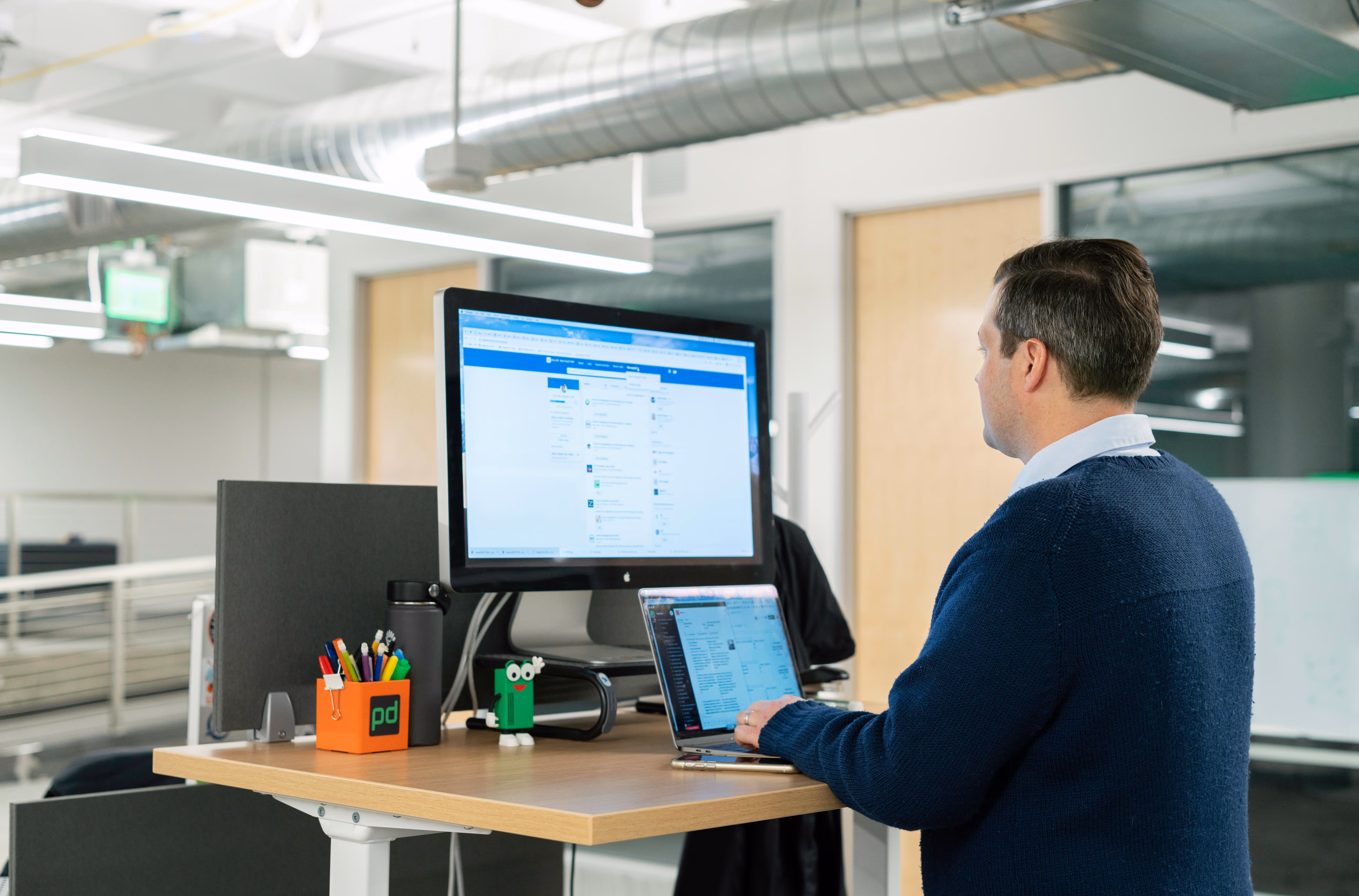Man in a navy sweater working on a laptop at a standing desk with a large external monitor in a modern office.