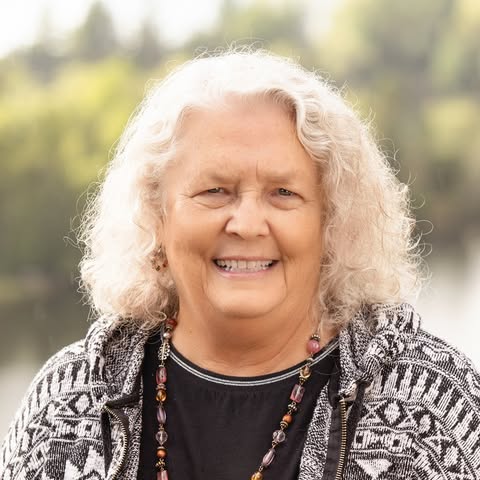 Headshot of Sharon, co-owner of Riverside Pet Lodge, a smiling woman with white curly hair, wearing a patterned black and white jacket and a beaded necklace.