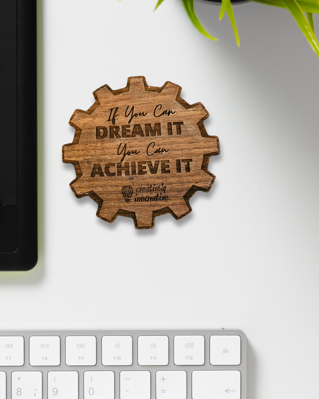 A top-down shot of a custom-engraved wooden coaster in a gear shape, with the text "If You Can DREAM It You Can ACHIEVE It" and the "Creatively Innovative" logo, resting on a clean white desk next to a black laptop and a keyboard.