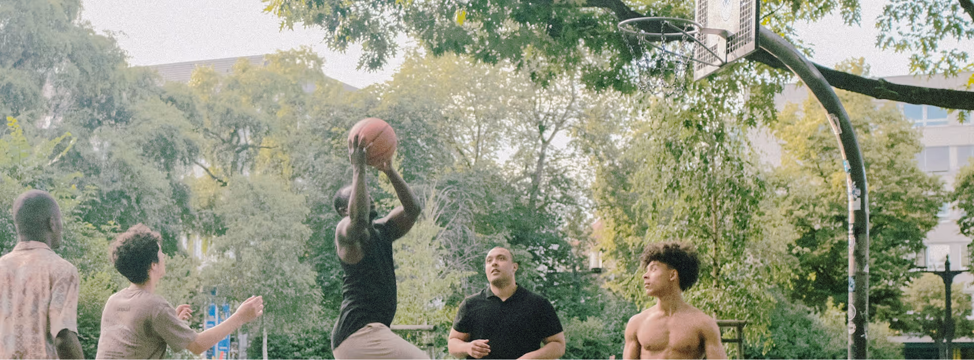 Group of young men playing basketball outdoors under trees, one jumping to shoot the ball toward the hoop.
