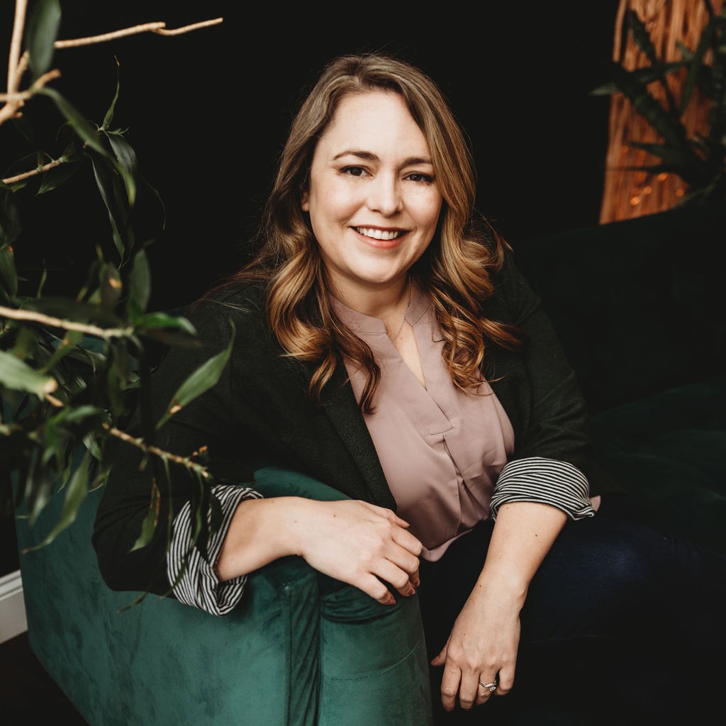 Smiling woman with wavy light brown hair sitting on a green velvet sofa, wearing a mauve blouse and dark blazer with striped cuffs against a dark background with indoor plants.