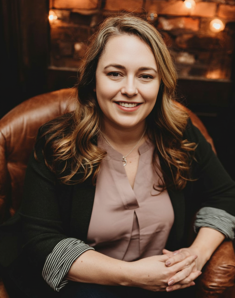 Smiling woman with wavy light brown hair sitting in a brown leather armchair, wearing a mauve blouse and dark blazer against a warm, brick wall background.