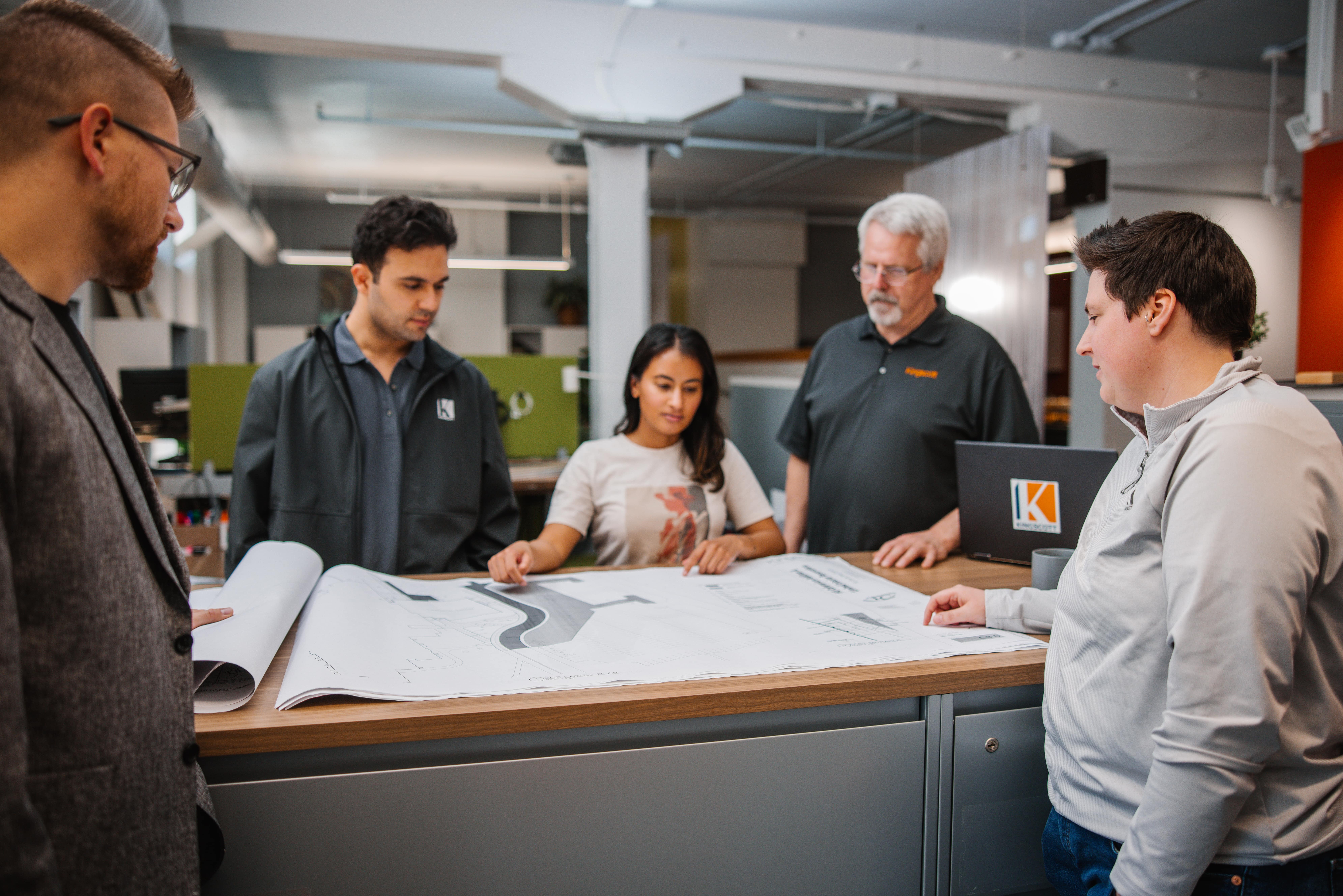 Five colleagues gathered around a table reviewing large architectural blueprints in an office.