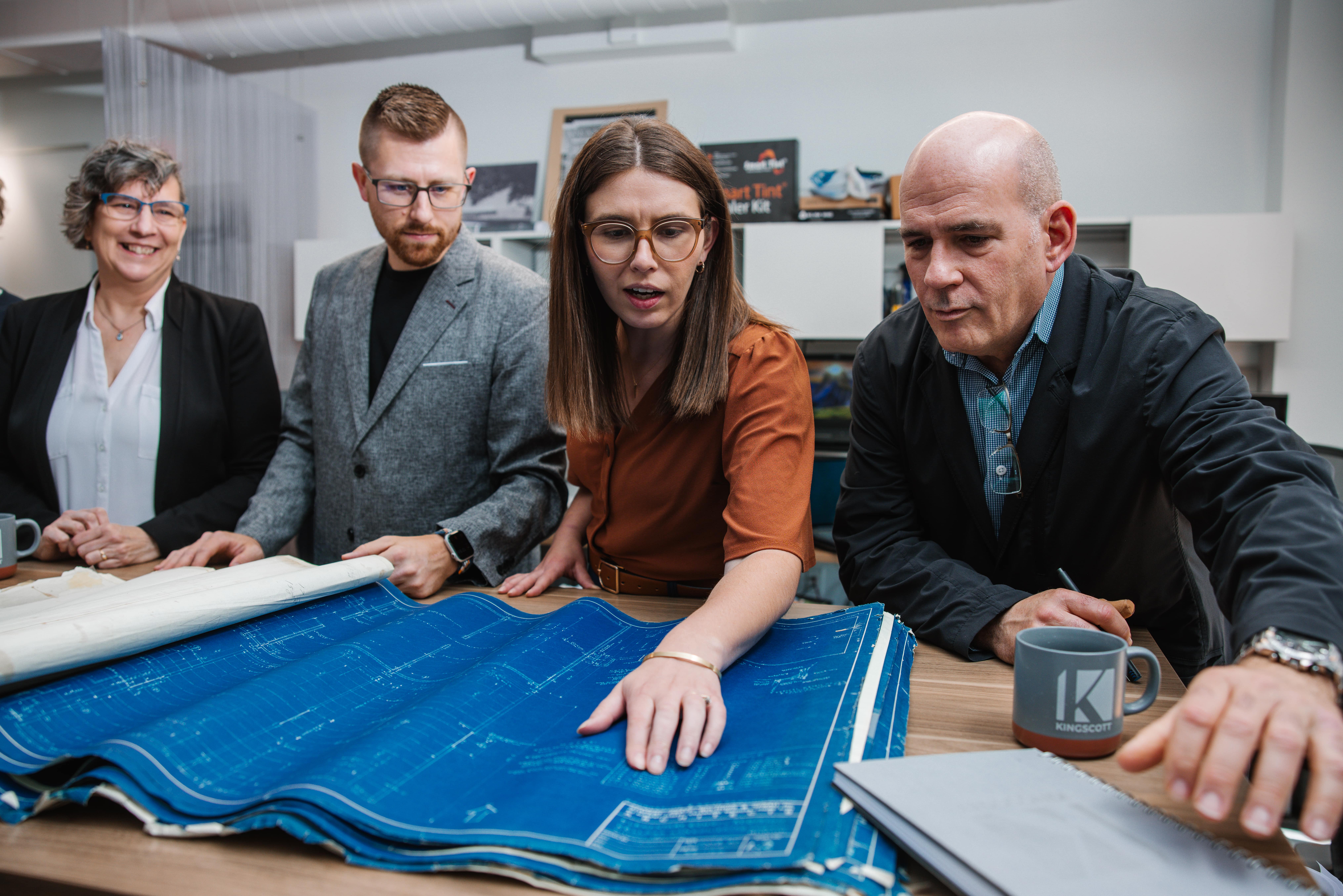 Four professionals examining blueprints spread on a desk in an office setting.