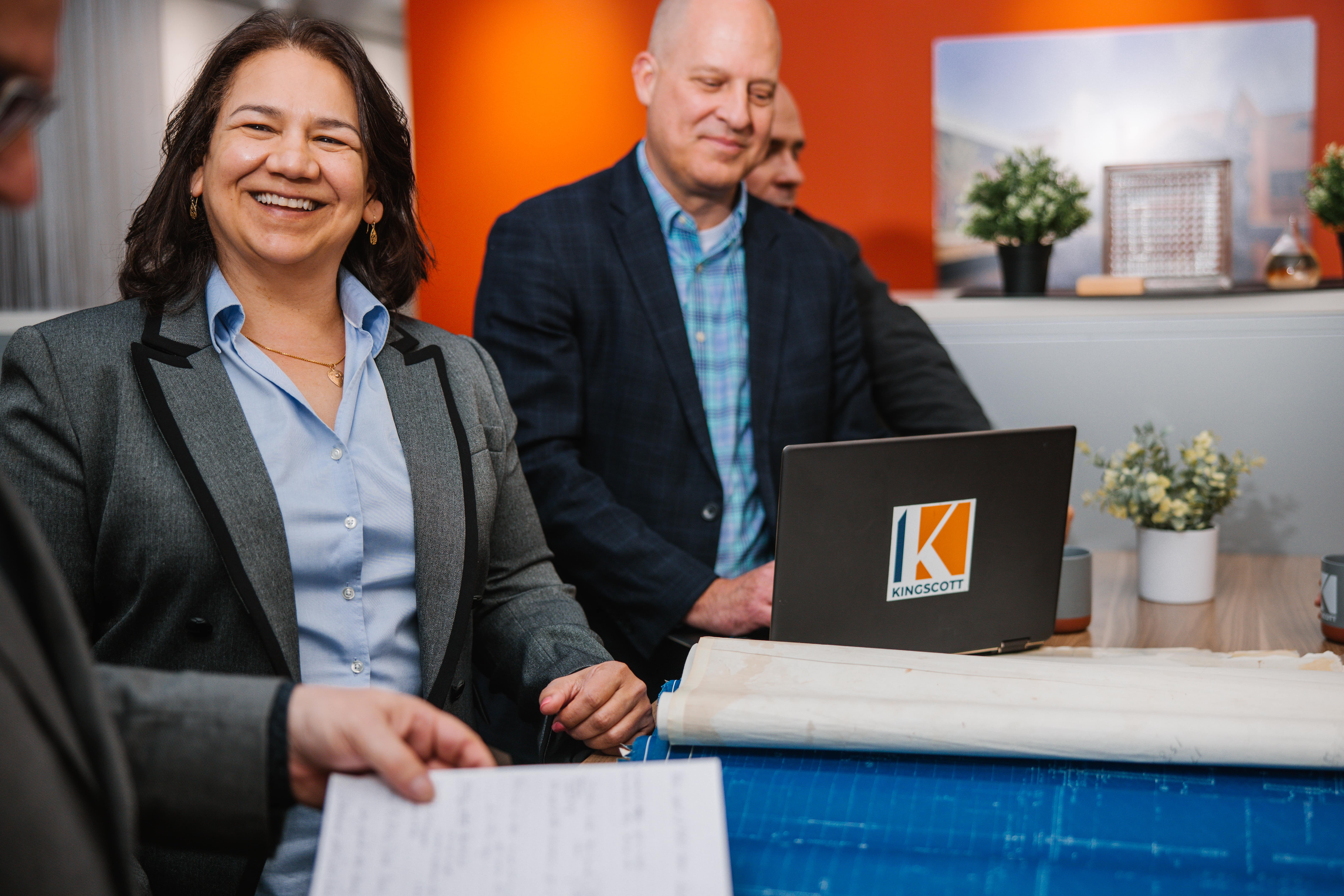Smiling woman in grey blazer and blue shirt sitting at a table with blueprints, two men behind using a laptop with Kingscott logo.