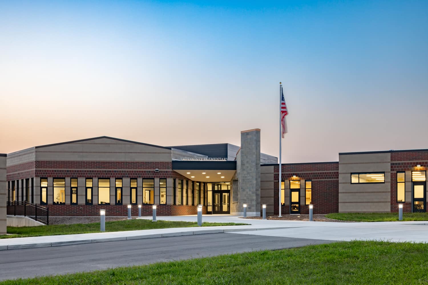 Modern elementary school building with brick and beige exterior, lit windows, and an American flag at dusk.