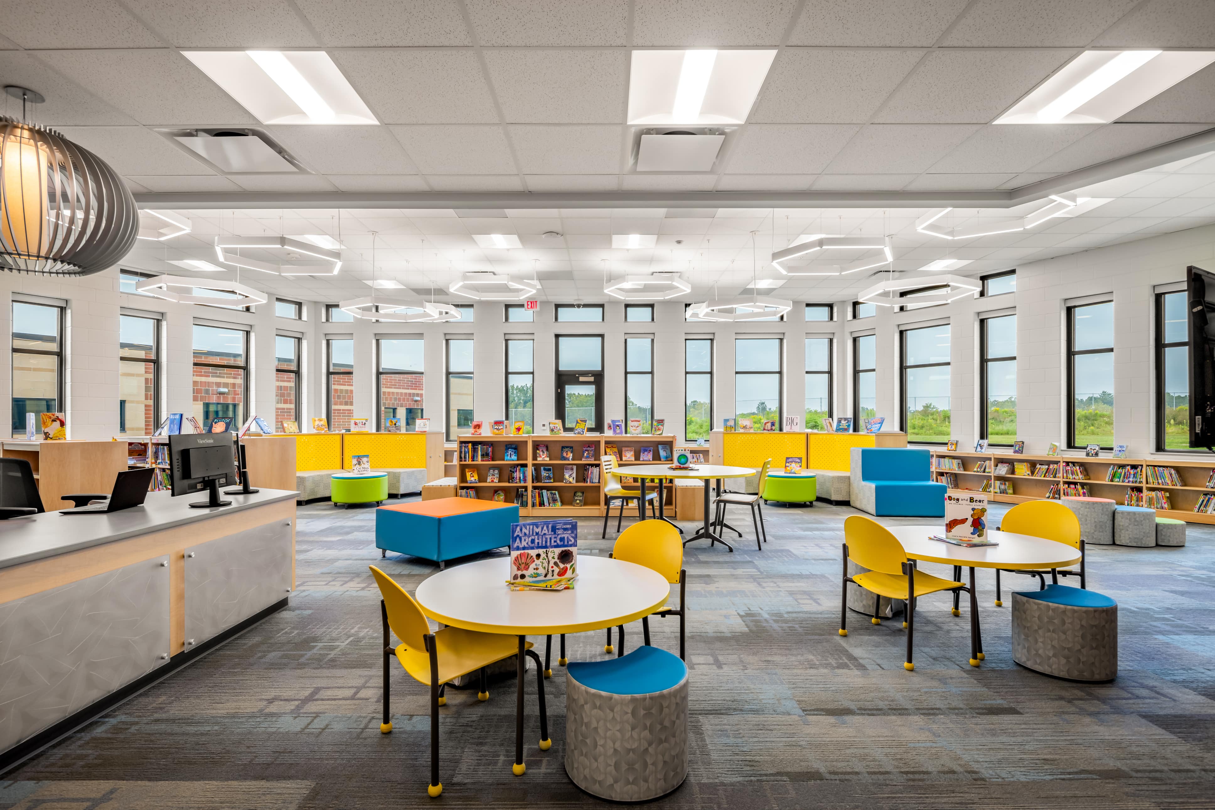 Bright, modern library with round white tables, yellow chairs, colorful seating cubes, bookshelves, and large windows.