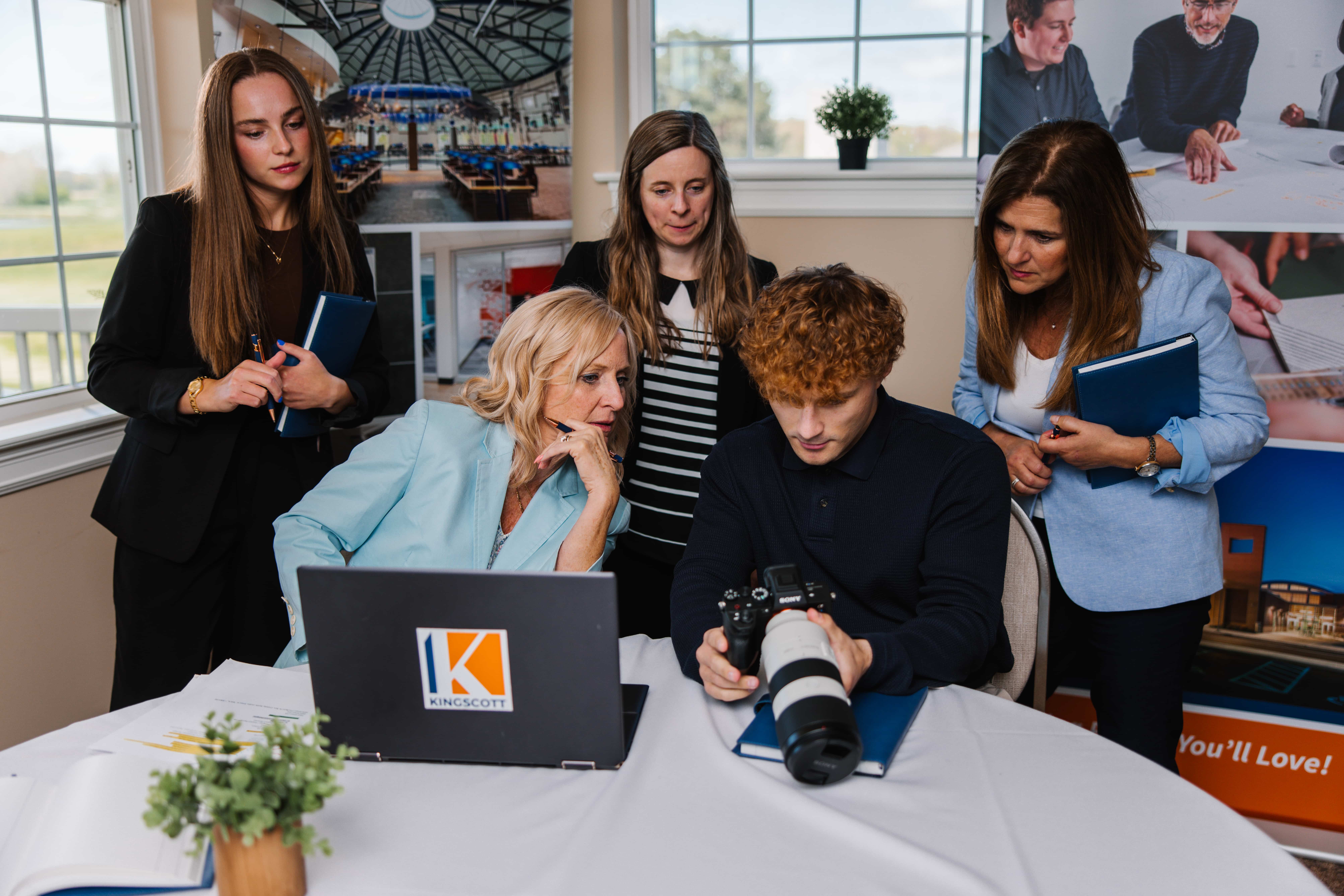 Five people gathered around a table, one reviewing photos on a camera while others watch, with a laptop displaying a Kingscott logo.