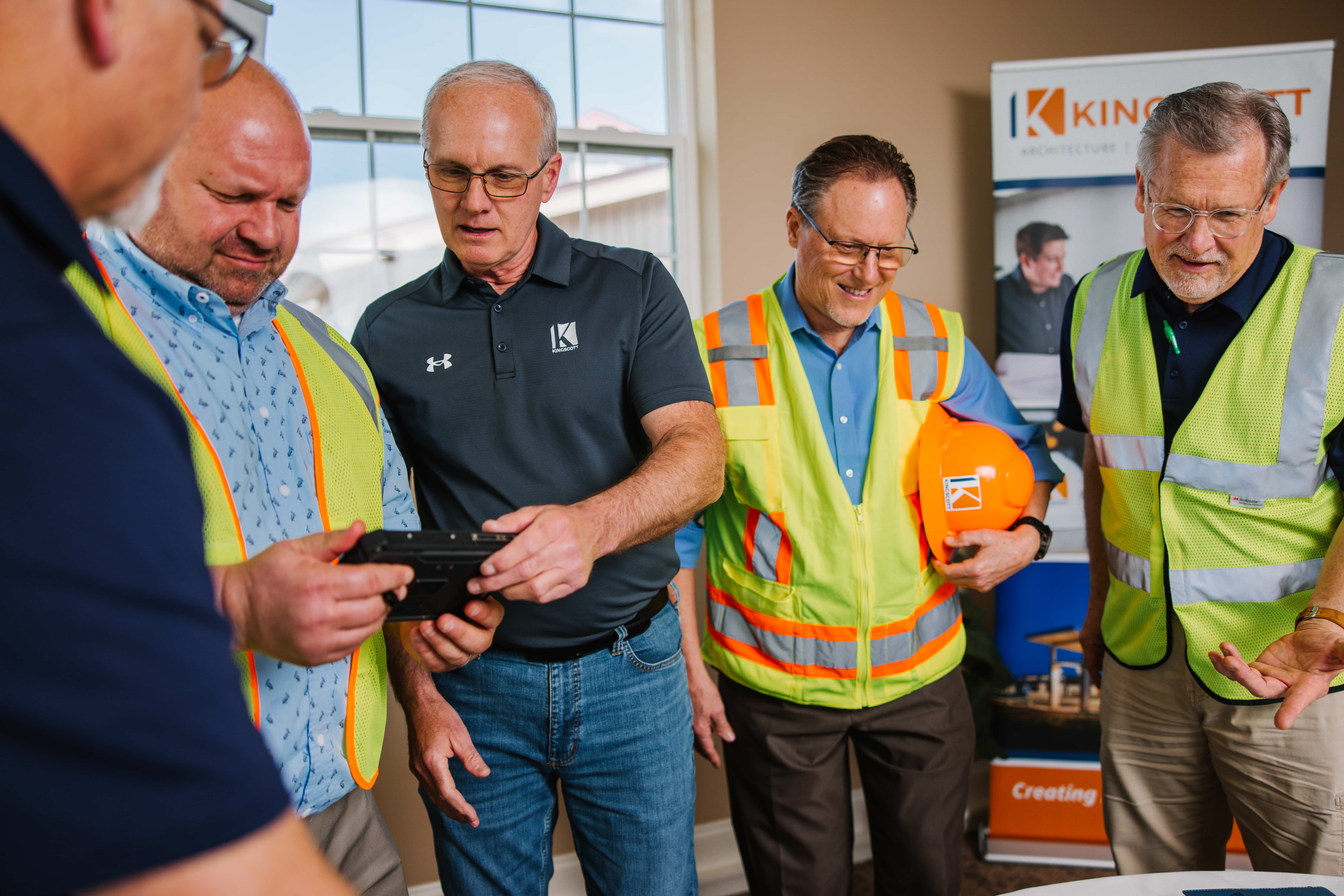 Five men, some wearing safety vests and holding a hard hat, gathered indoors looking at a device held by one man.