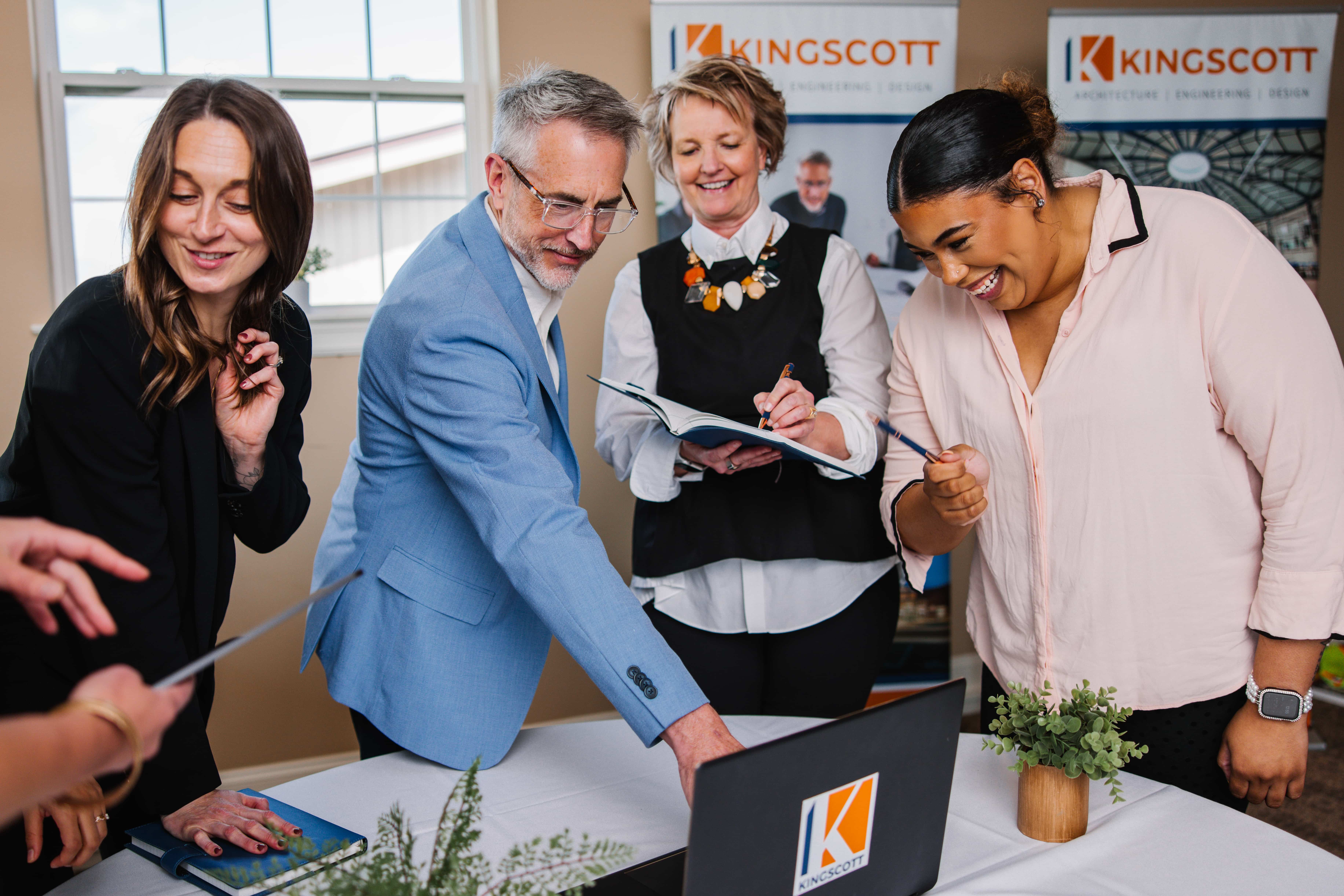 Four colleagues smiling and collaborating around a table with a laptop and notebook in an office setting.