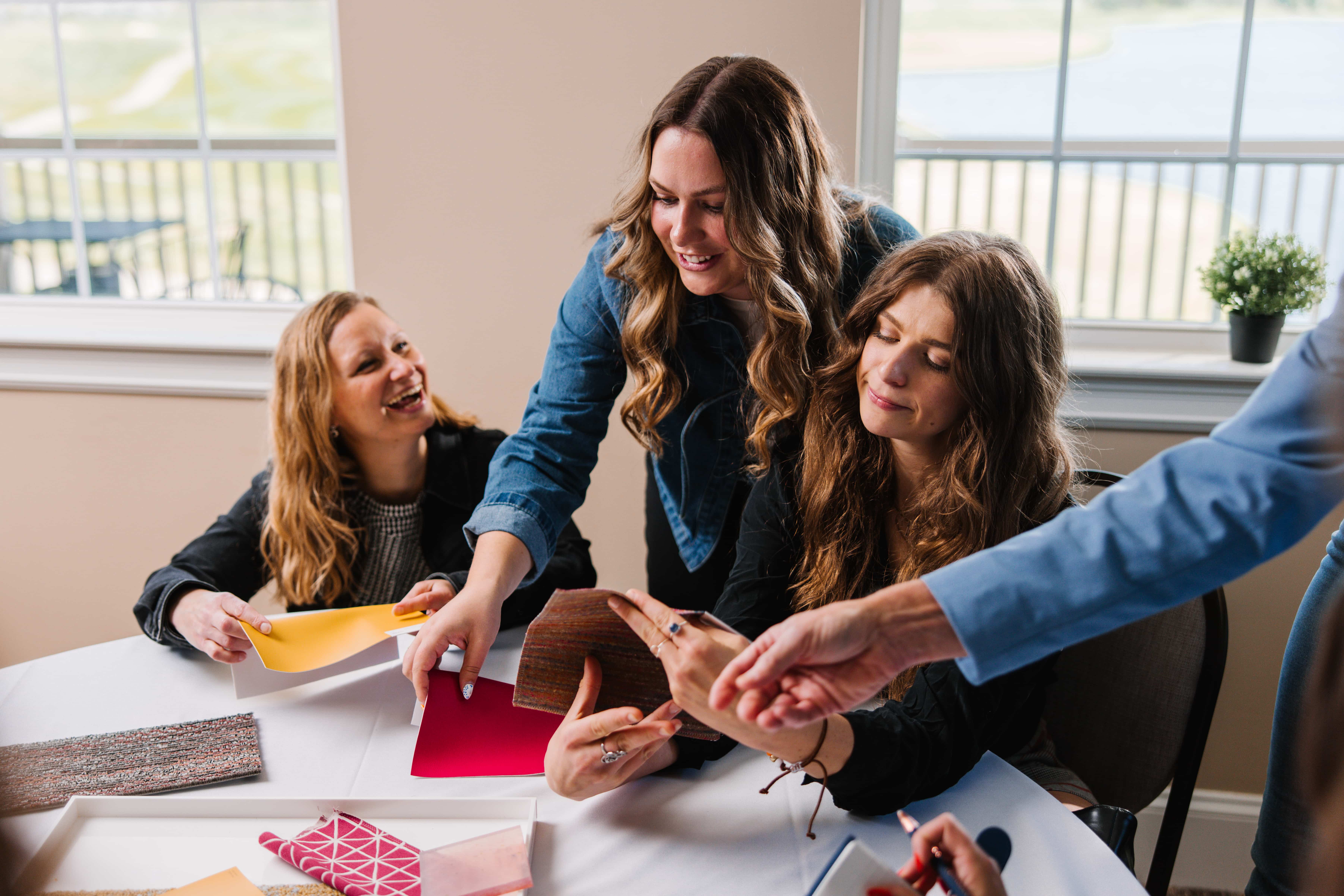 Three women reviewing various color and fabric samples around a table in a well-lit room with windows.