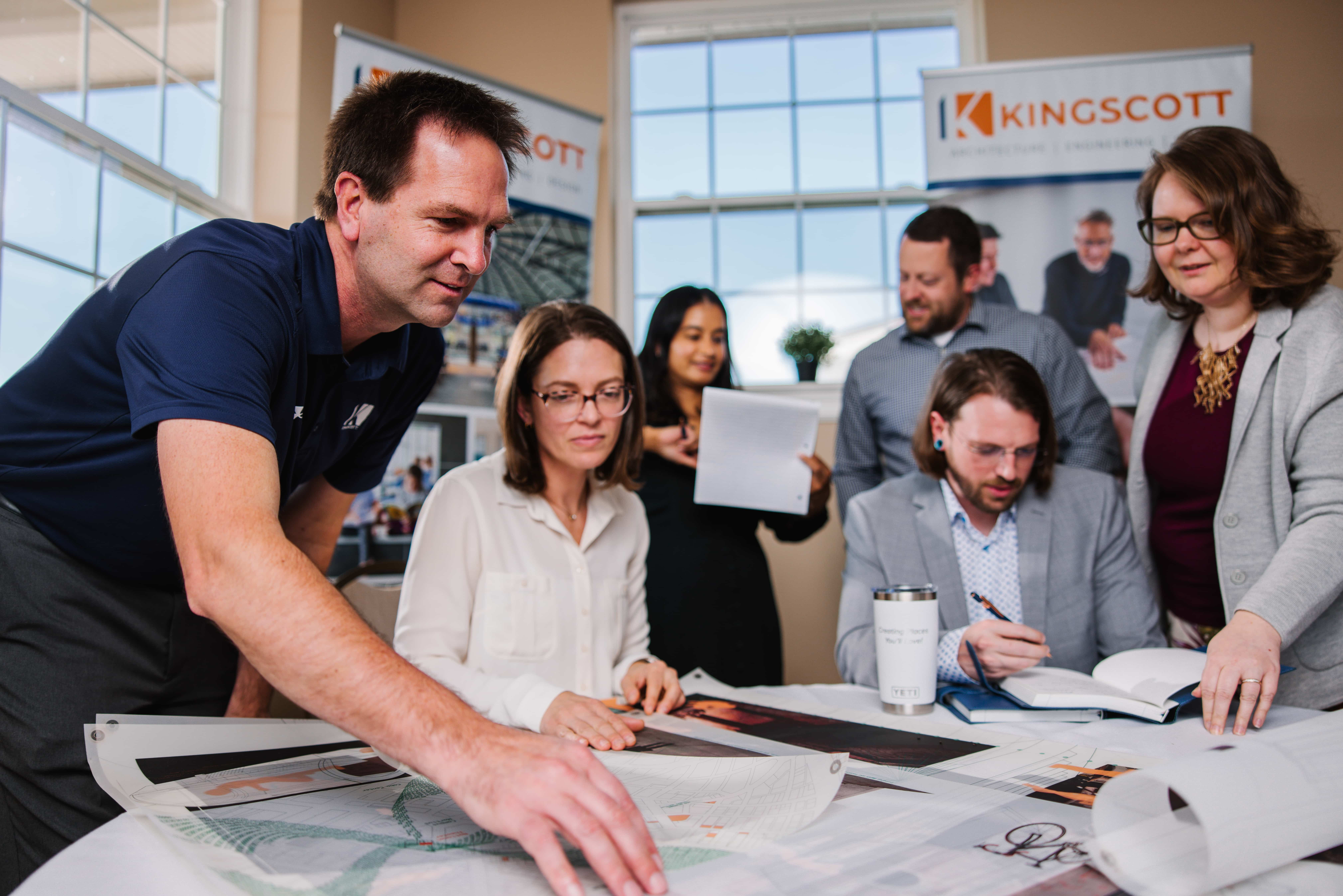 A group of six professionals collaborating over architectural plans spread on a table in a bright office with Kingscott banners in the background.