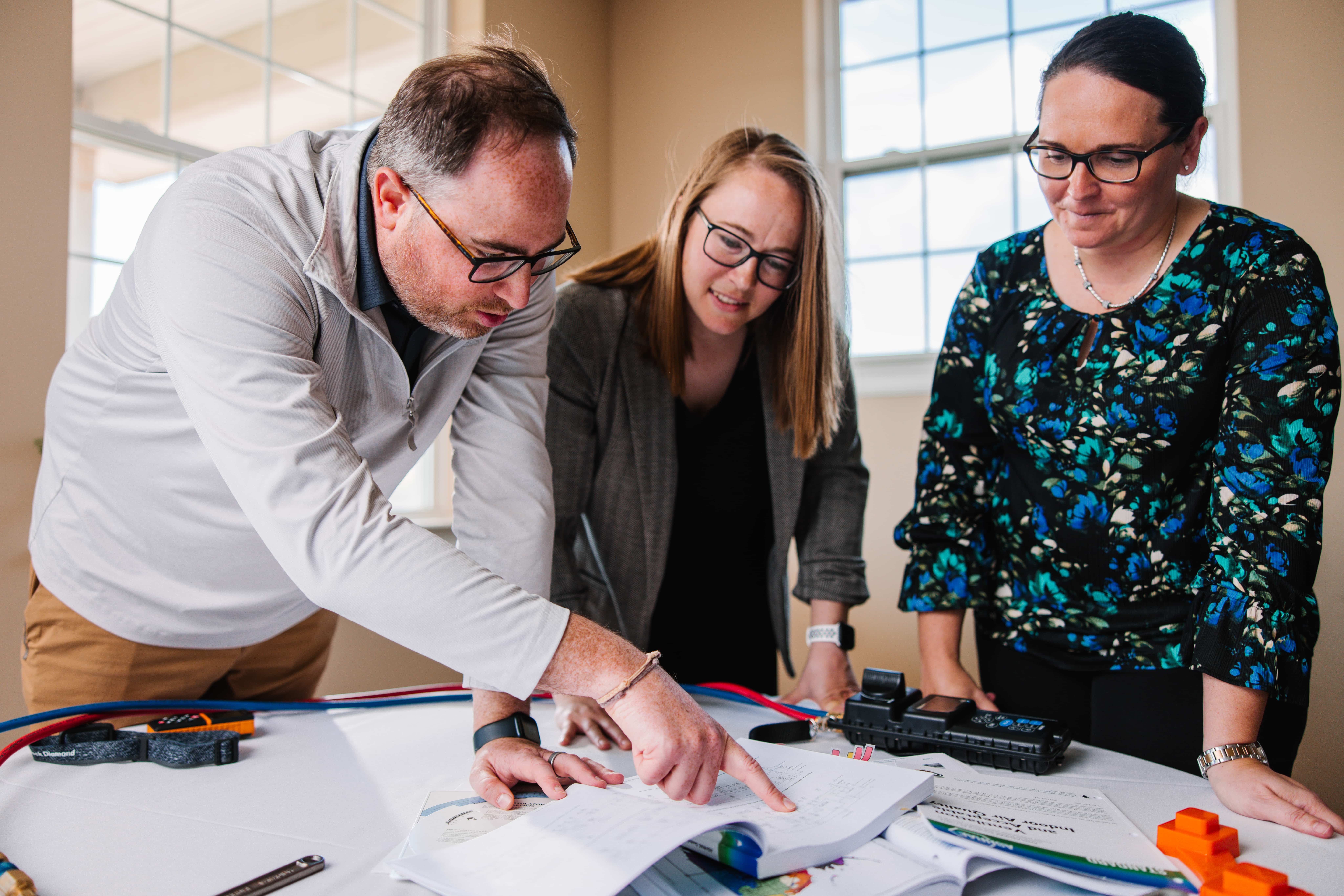 Three people wearing glasses reviewing documents and technical equipment on a table in a room with large windows.