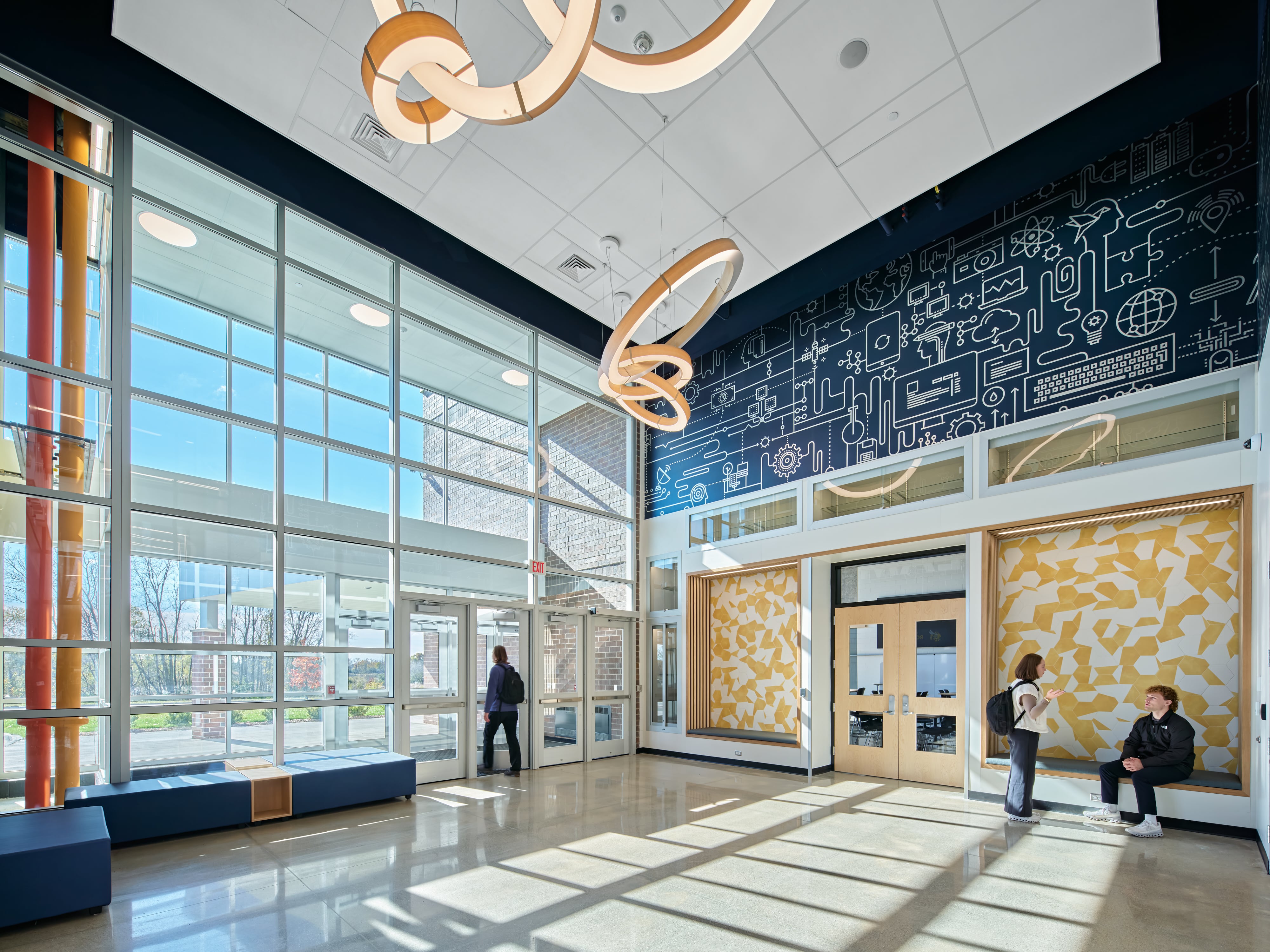 Bright, modern library with round white tables, yellow chairs, colorful seating cubes, bookshelves, and large windows.