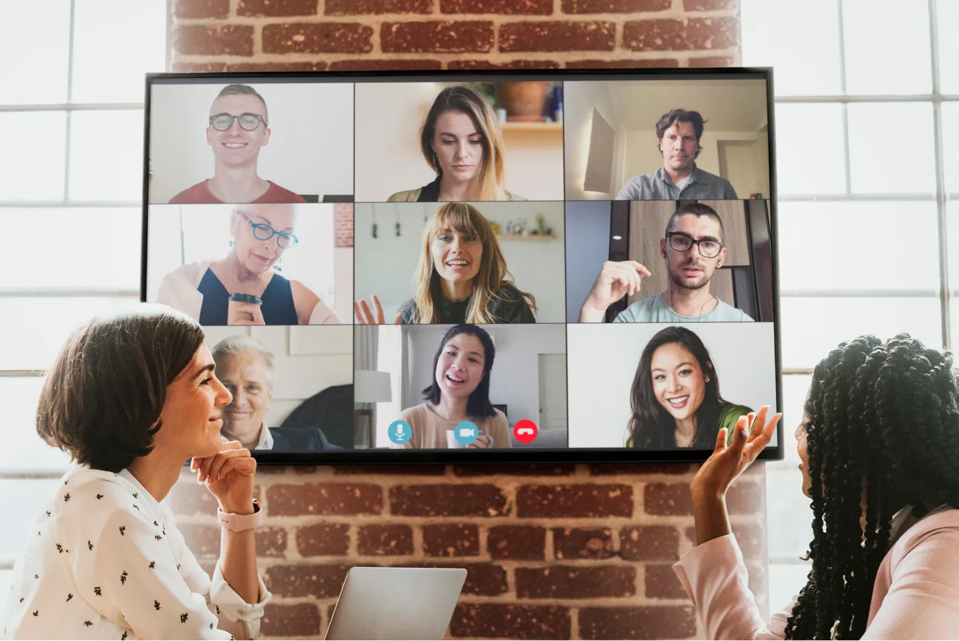 Two women sitting at a table video conferencing with nine colleagues displayed in a grid on a wall-mounted screen.