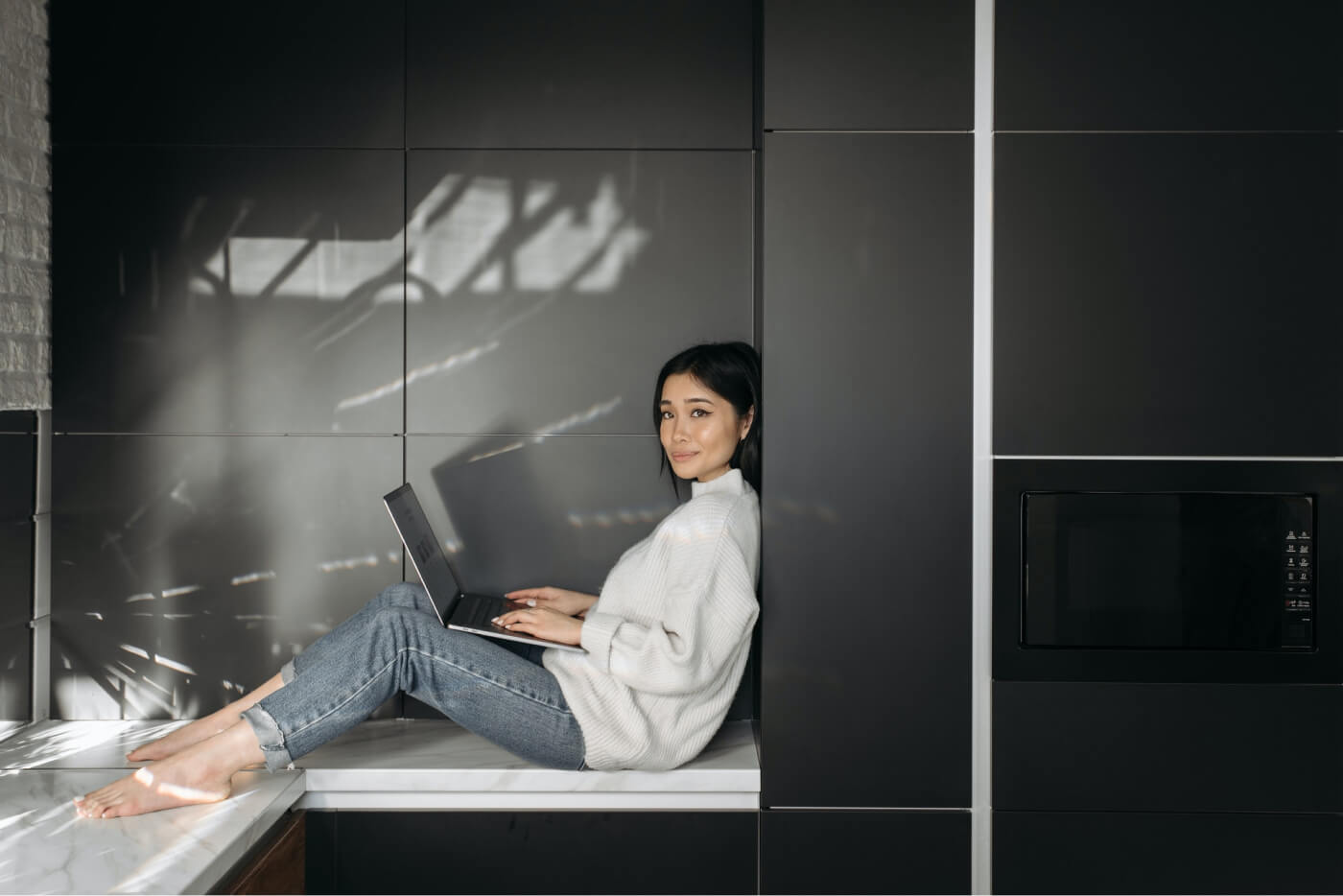 Woman sitting barefoot on a kitchen counter with a laptop, leaning against black cabinets.
