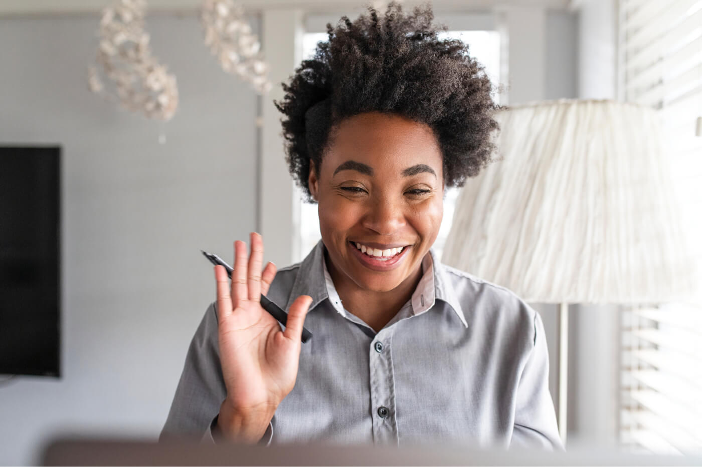 Smiling woman in a gray shirt waving and holding a pen during a video call.