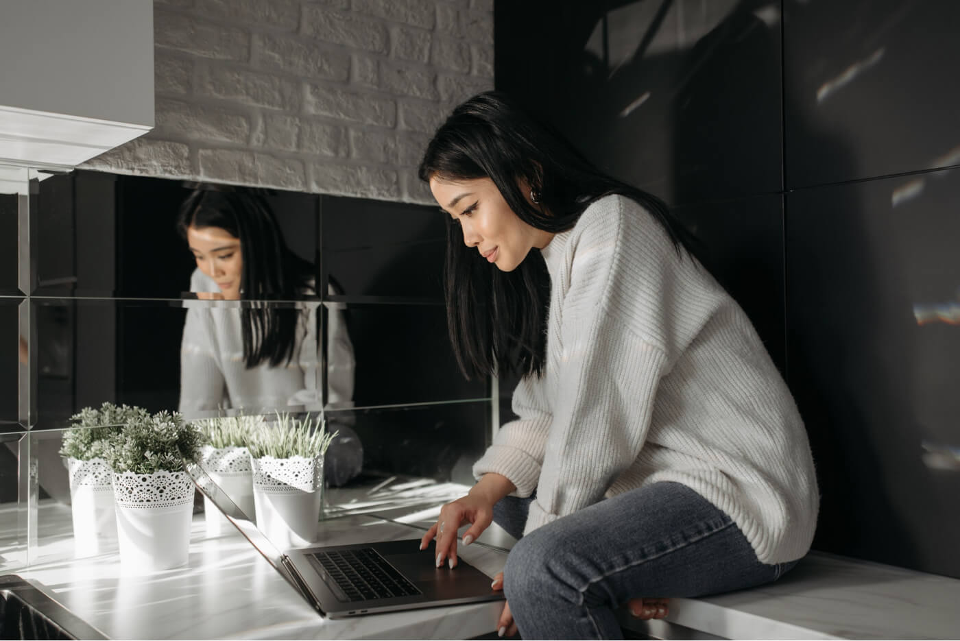 Woman in white sweater sitting on a marble countertop using a laptop next to three potted green plants and a mirror wall.