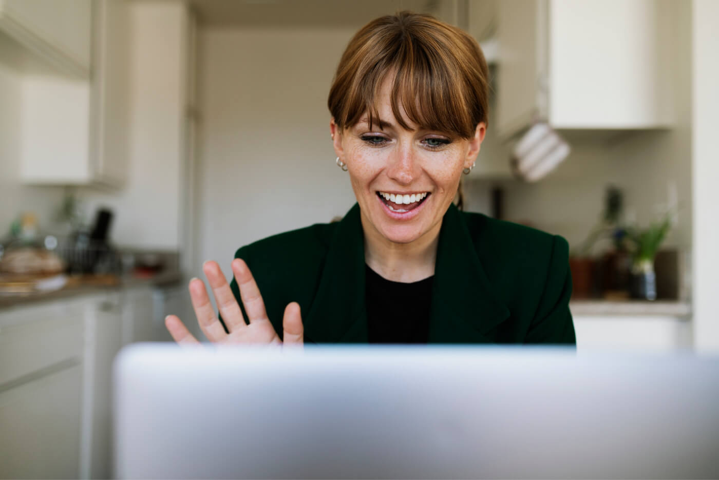 Smiling woman waving during a video call at a laptop in a kitchen.