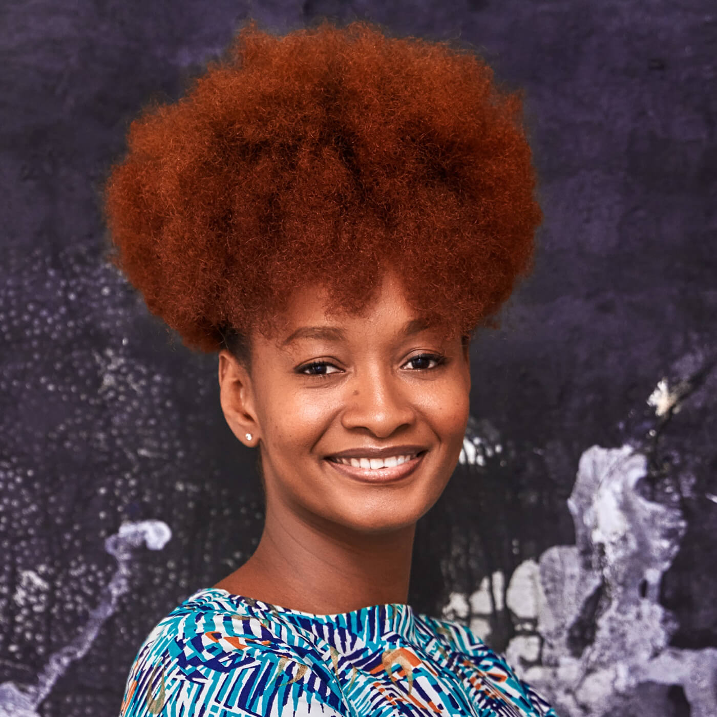 Smiling woman with vibrant red curly hair wearing a colorful patterned top standing in front of a dark abstract background.