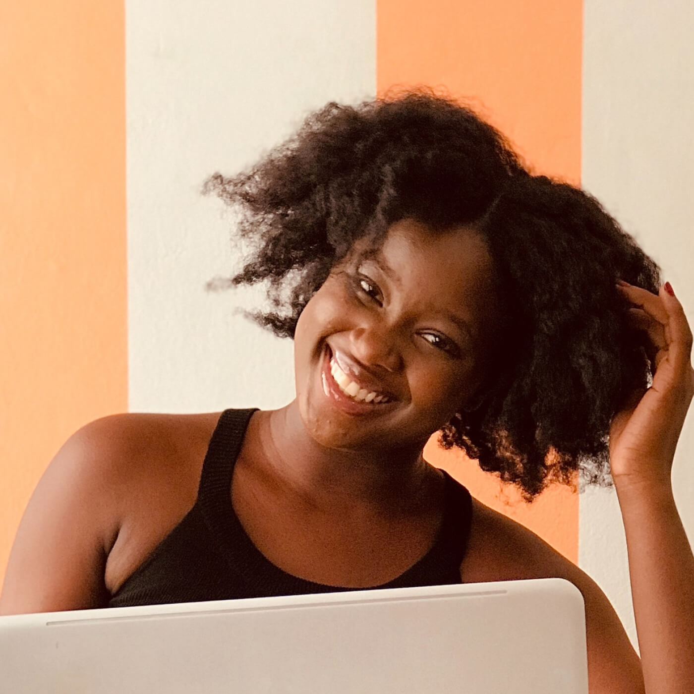 Smiling woman with natural curly hair wearing a black tank top, sitting in front of a white laptop with an orange and white striped background.