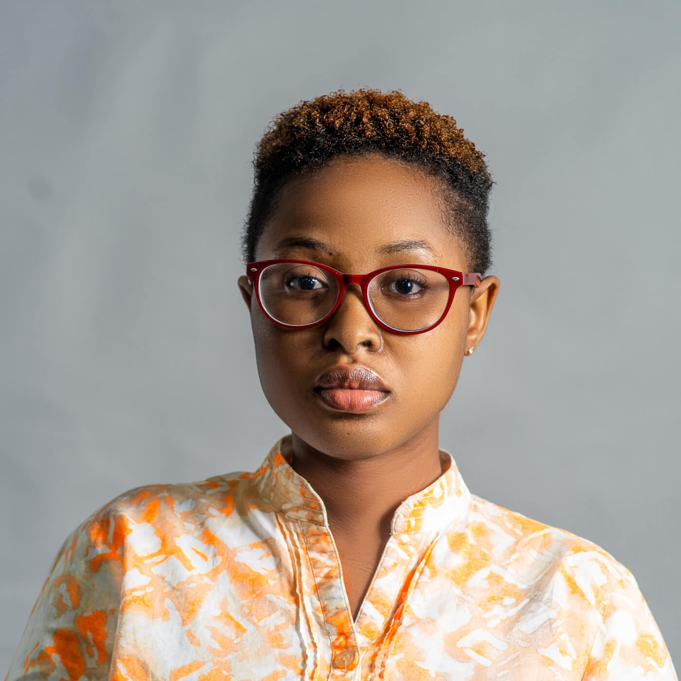 Portrait of a person with short curly hair, wearing red glasses and a white and orange patterned shirt, against a plain gray background.