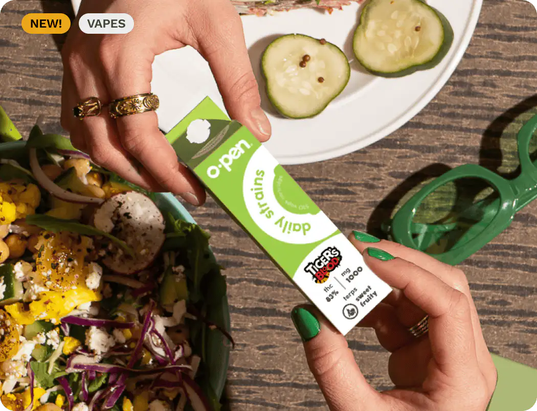 Two hands holding a green and white vape product package labeled daily strains and Tiger's Blood, with green sunglasses and plates of salad and cucumber slices in the background.